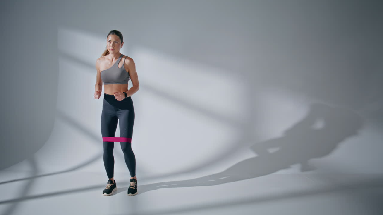 Female athlete doing squats with elastic band on white wall background indoors