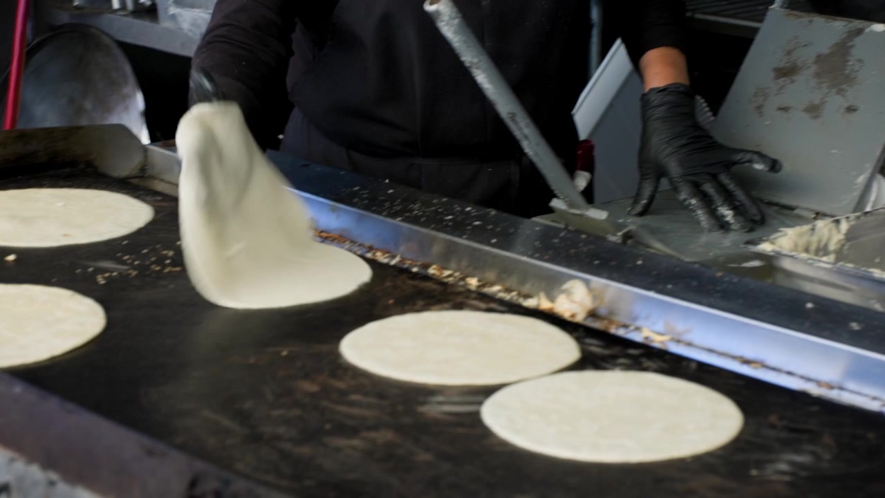 Filmagem de culinária tradicional mostrando uma senhora ao fundo fazendo tortilhas com uma chapa em primeiro plano durante a preparação de tortilhas de milho frescas e artesanais