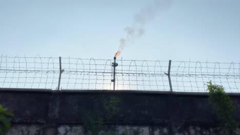 Gasfackel-Industrieanlage-Hinter-Einer-Schutzmauer-Aus-Drahtzaun,-Sonnenuntergang