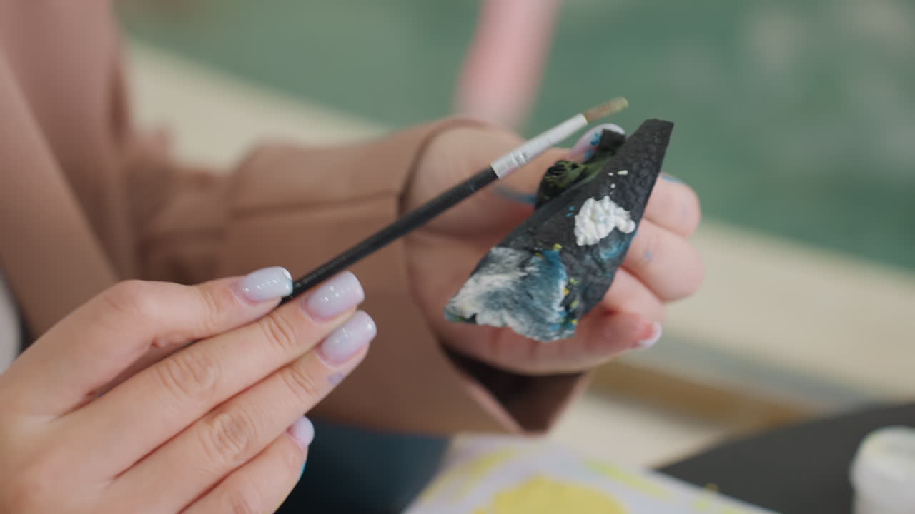 primer plano de manos blancas femeninas pintando detalles blancos en un modelo de barco en miniatura negro con un pincel fino, fondo de estudio exterior desenfocado, trazos cuidadosos y pequeños botes de pintura visibles, calma artesanal