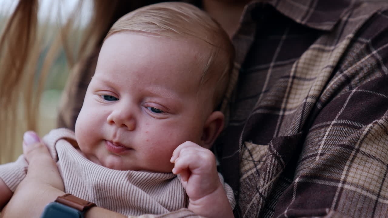 Cute sleepy Caucasian infant boy with blond hair and blue eyes. Close up. Little baby in hand of parents standing in the nature.