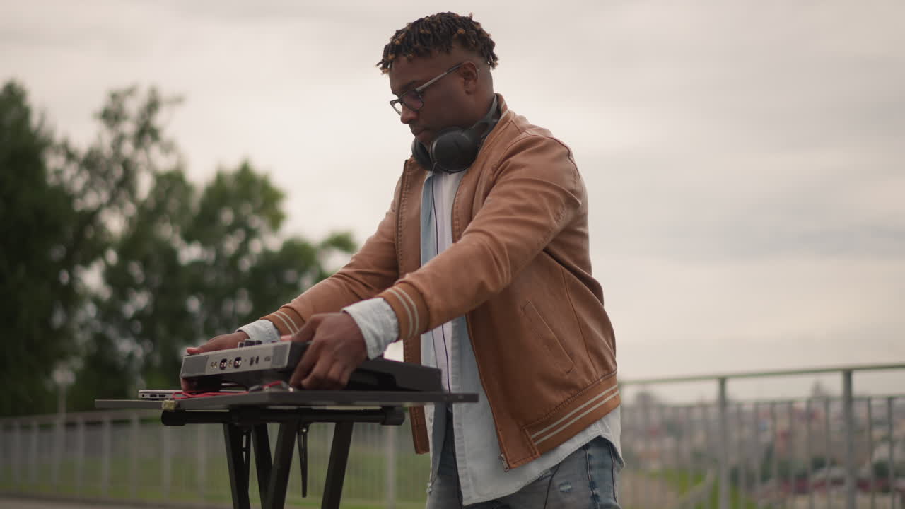 dj on rooftop mixing soulful tracks, wearing brown jacket and headphones, using laptop and controller on portable stand near railing, overcast sky and park backdrop, focused expression adjusting knobs