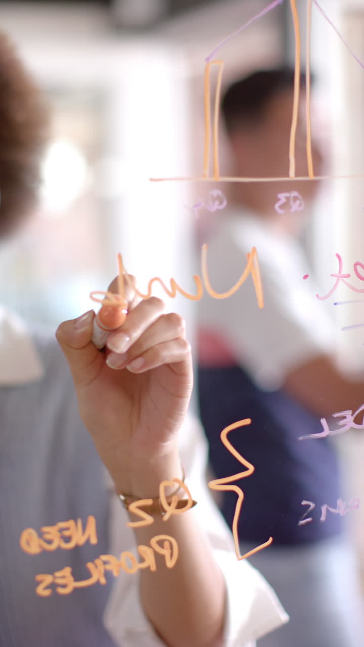 Vertical video of thoughtful biracial businesswoman brainstorming on office glass wall, slow motion