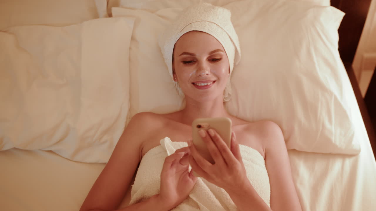 mujer relajándose en la cama con una máscara facial y usando un teléfono inteligente