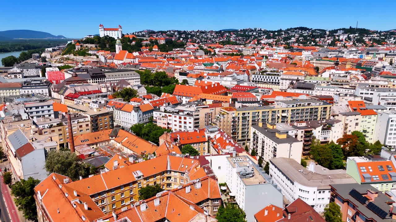 Flying over the beautiful historical buildings with orange roofs. Bratislava Castle on the green hill at backdrop