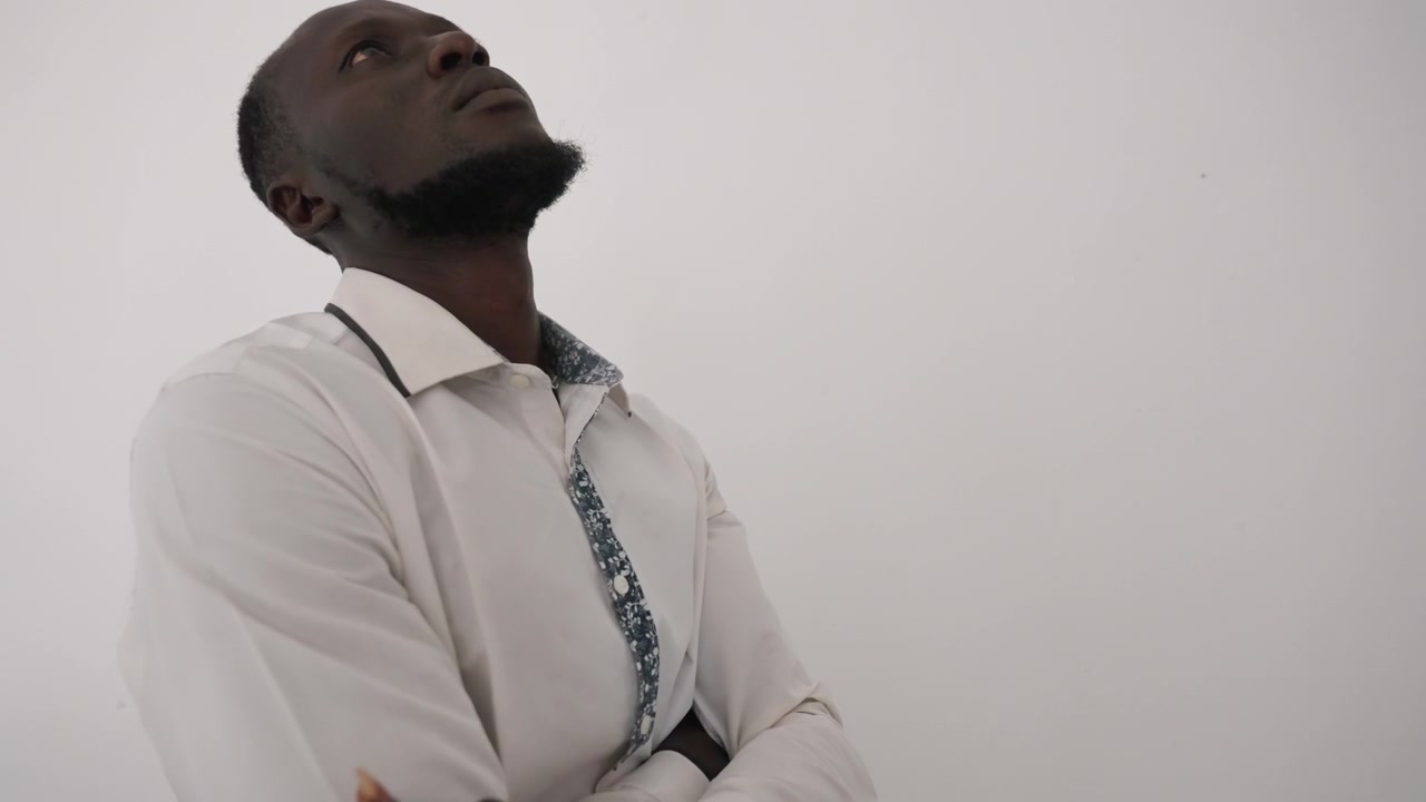 Uomo di colore in camicia bianca, dall'aria speranzosa, che guarda in alto immaginando una serata romantica, corteggiatore paziente in attesa dell'appuntamento, leggera inclinazione del petto, braccia incrociate, sfondo minimale da studio, umore da sogno tranquillo.