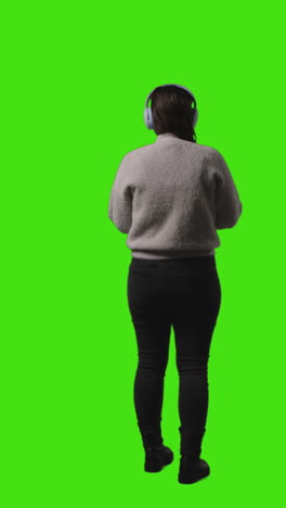 vertical video rear view full length shot of woman wearing wireless headphones streaming music from mobile phone against green screen 2