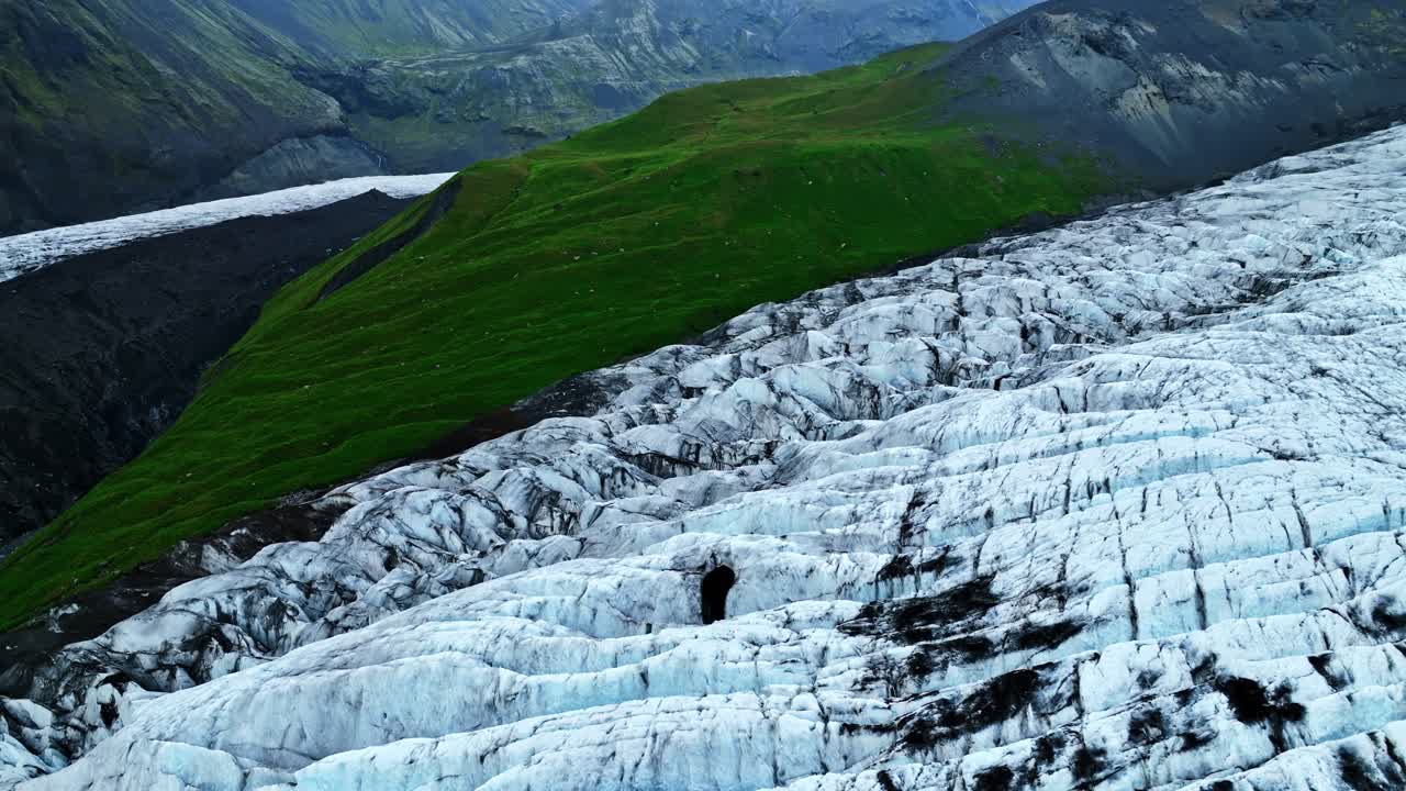 Aerial shot of a rugged Icelandic glacier flowing beside a vivid green mountain ridge, revealing sharp contrasts between ice, moss-covered slopes, and dramatic volcanic terrain