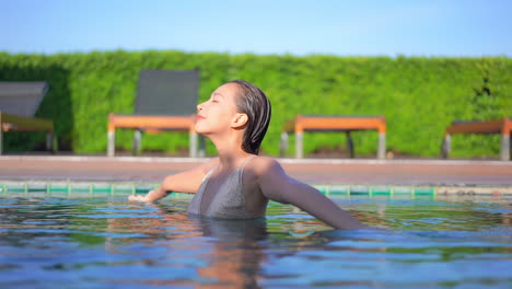 asian woman in swimwear turns around with spread on sides hands and closed eyes inside swimming pool at a private resort in spain, slow-motion daytime sunny