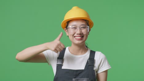 close up of asian woman worker wearing goggles and safety helmet smiling and showing thumbs up gesture while standing in the green screen background studio