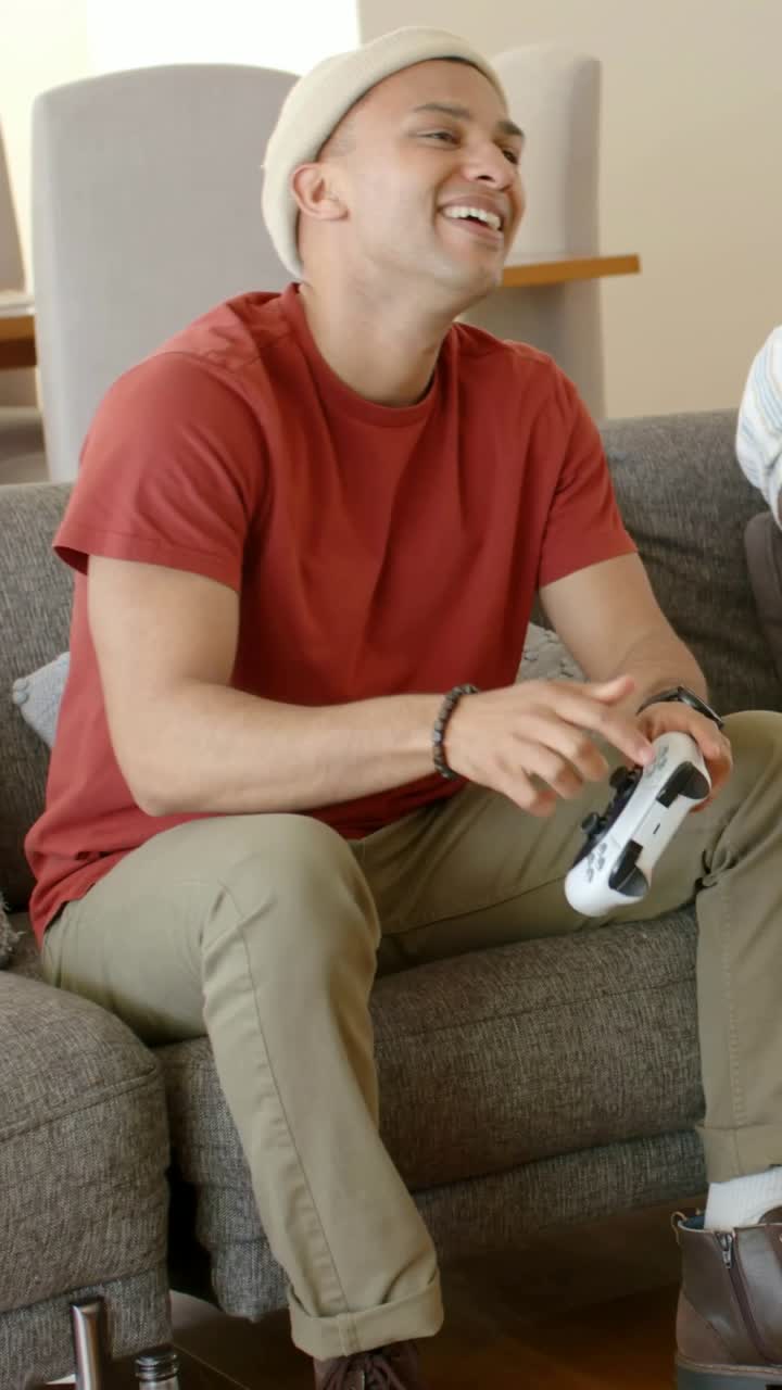 Vertical video: Leaning for bottle, man in beanie grabbing gamepad in living room, playing together
