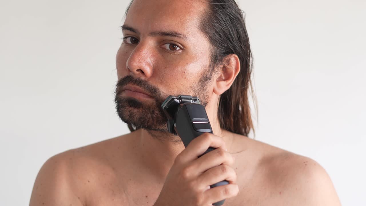 hombre latino de cabello largo afeitando con afeitadora eléctrica barba y bigote en el baño, espejo
