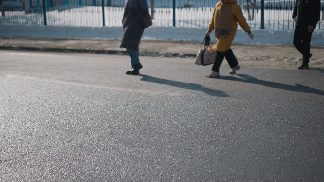 lower view pedestrians in coats and boots walking across pavement, long shadows stretching over wet asphalt, people carrying bag and backpack during cold winter commute beside snowbank