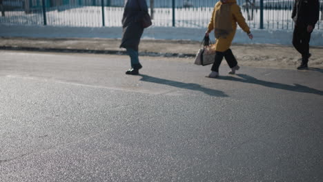 lower view pedestrians in coats and boots walking across pavement, long shadows stretching over wet asphalt, people carrying bag and backpack during cold winter commute beside snowbank