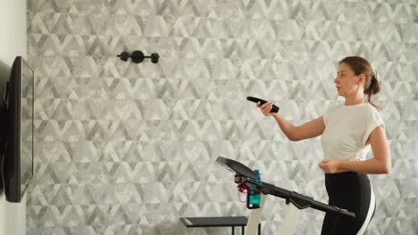 woman turns tv channels training at home. graceful lady switches on television running on treadmill in house. woman watches television at jogging indoors