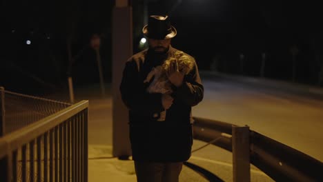 man at night in a trench coat walking along a bridge holding a baby doll