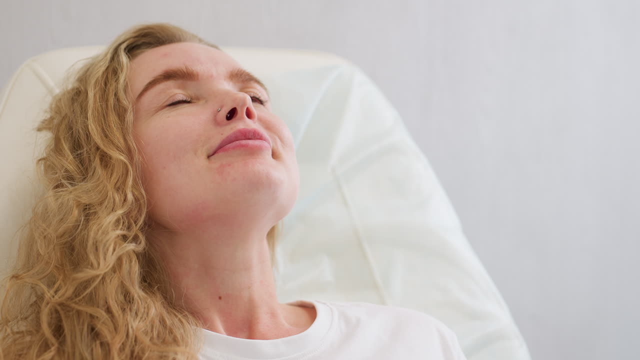 Blond client wearing white polo shirt gently smiles while lying back on chair during beauty or wellness session, conveying calm, relaxation, and personal care in soft-lit setting