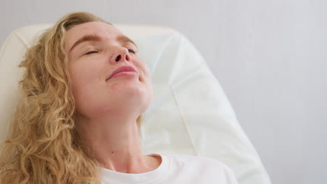 blond client wearing white polo shirt gently smiles while lying back on chair during beauty or wellness session, conveying calm, relaxation, and personal care in soft-lit setting