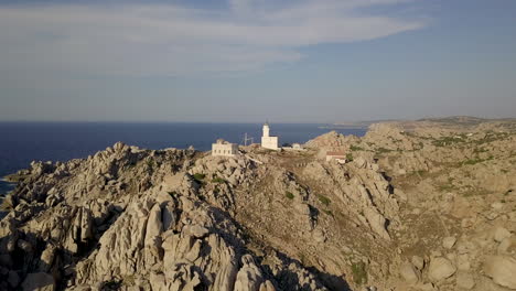 Drone-shot-rising-up-along-a-rocky-cliff-in-Sardinia-with-a-white-light-house-on-it-to-reveal-a-pristine-bay-behind-it