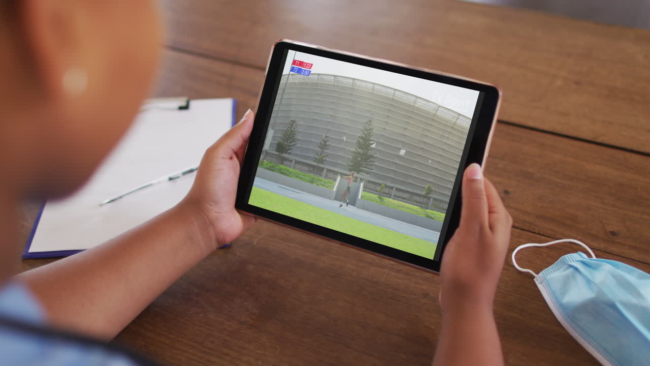 태블릿에서 육상 자벨린 경기를 시청하는 얼굴 마스크와 함께 테이블에 앉아있는 여성의 복합