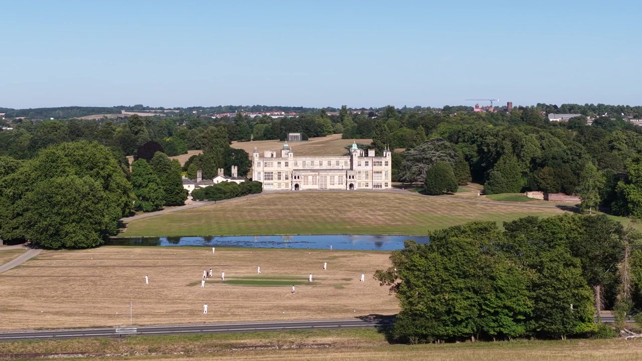 Splendida vista panoramica di una tradizionale partita di cricket inglese che si svolge su un campo bruciato dal sole, con la maestosa villa di Ashton Court a fare da sfondo storico in una limpida giornata estiva.
