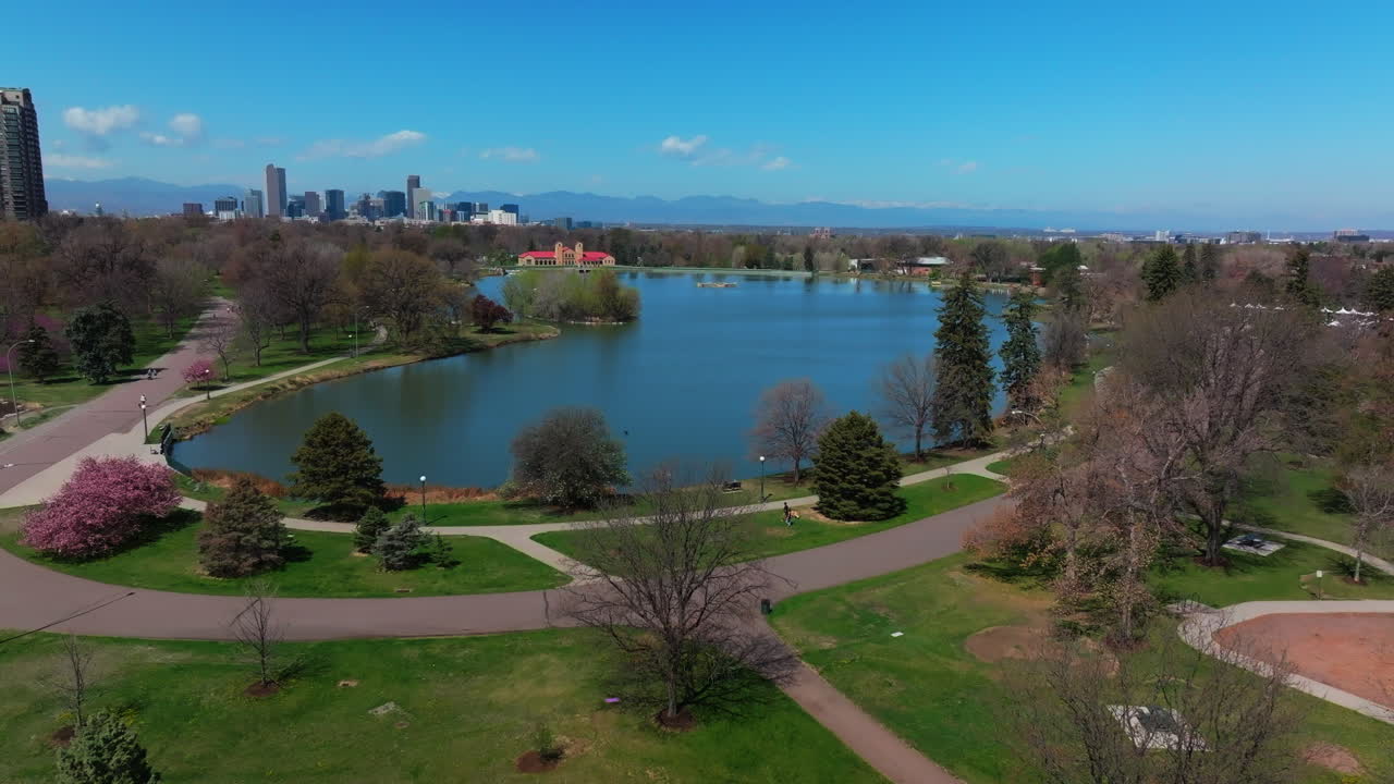 City Park Denver Colorado Lake Pavilion vibrant spring summer aerial drone sunny blue sky snow capped Rocky Mountains front range cityscape green lush grass trees blossom forward upwards motion