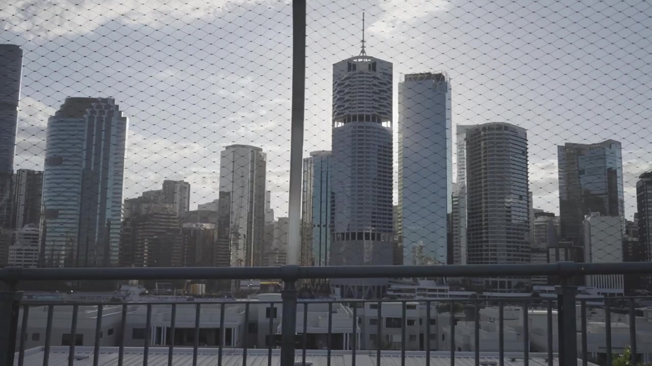 O horizonte da cidade de Brisbane é visto através de uma cerca de segurança de malha com padrão de diamante em um mirante elevado