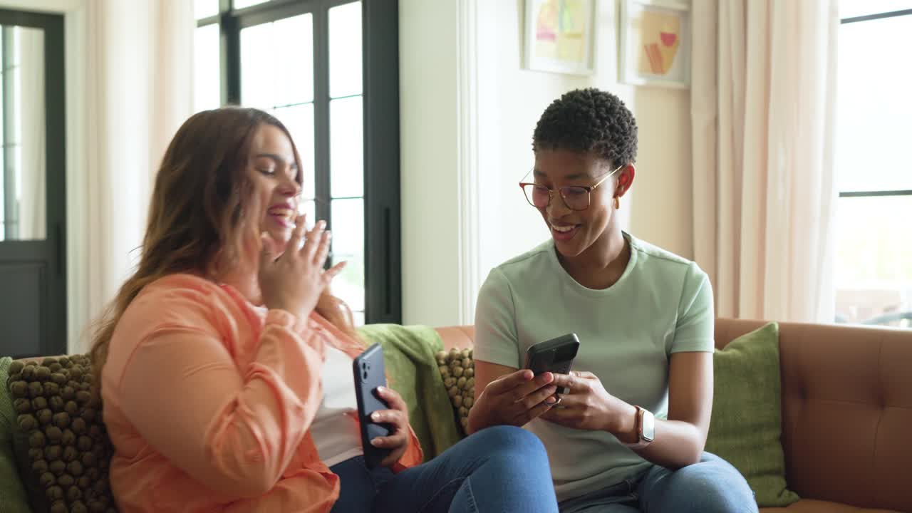 Diverse women on sofa, left woman scrolling phone, sparking right woman checking phone, laughing