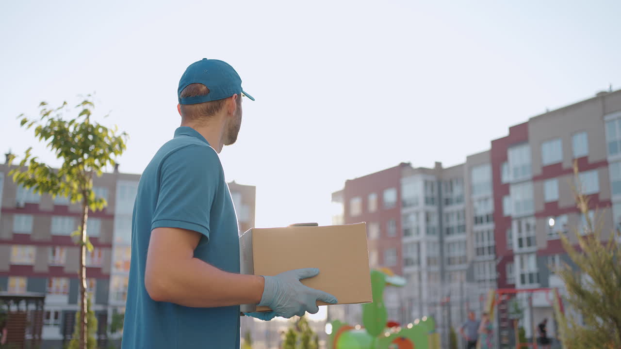 A delivery man in a blue T-shirt and cap carries a cardboard box in the city looking around in search of the deliveryman's address. Deliveryman goes with a box in slow motion