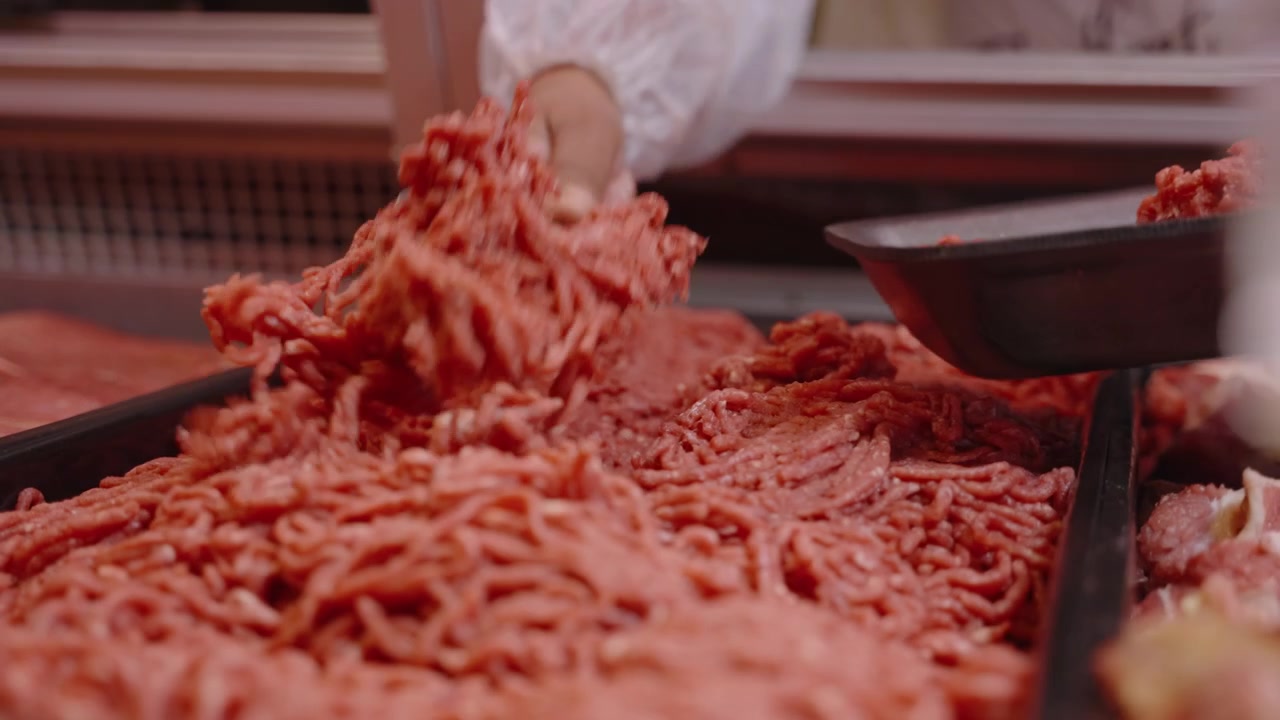 Gloved worker uses a metal scoop to portion raw minced beef from a chilled case at a retail butchery.