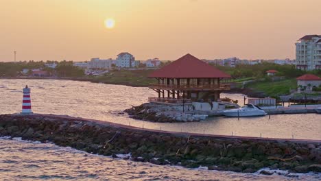 Toma-Aérea-Amplia-Que-Muestra-El-Agua-Del-Mar-Chocando-Contra-El-Embarcadero-Con-El-Faro-En-El-Puerto-Deportivo-De-La-Romana-Durante-La-Puesta-De-Sol---Hotel-De-Lujo-Hilton-Garden-Inn-En-Segundo-Plano