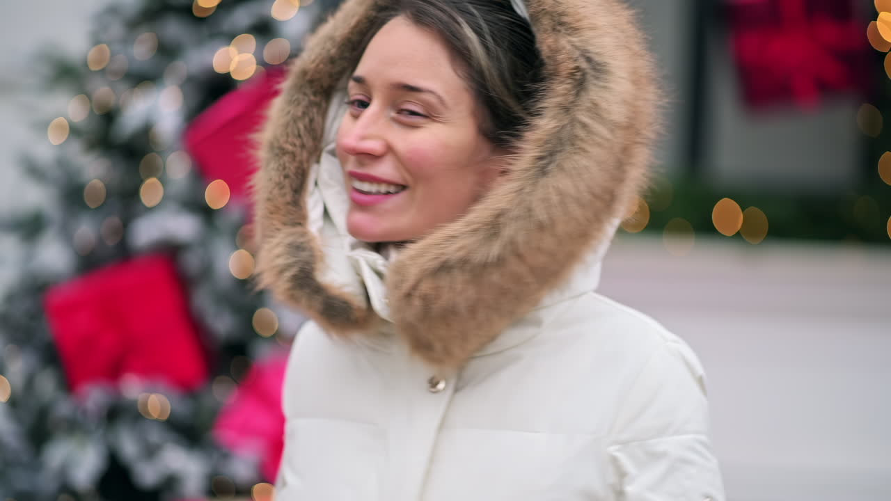 A woman smiles while wearing a cozy coat with a fur hood, surrounded by twinkling lights and holiday decorations in a vibrant city square, capturing the festive spirit