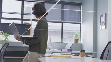 Biracial-businesswoman-standing-at-table-and-using-laptop-alone-at-office