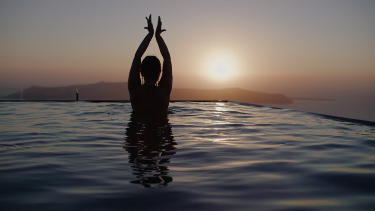 Una donna che balla in una piscina a sfioro guardando il tramonto sul mare - Rallentatore, Santorini, Grecia