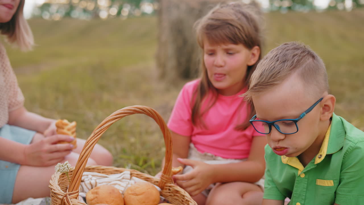 Un bambino con la maglietta verde si riempie la bocca con tutto il suo spuntino mentre sua sorella con la maglietta rosa si gusta il suo più delicatamente, con la mamma che osserva all'aperto in un campo durante un picnic in famiglia.