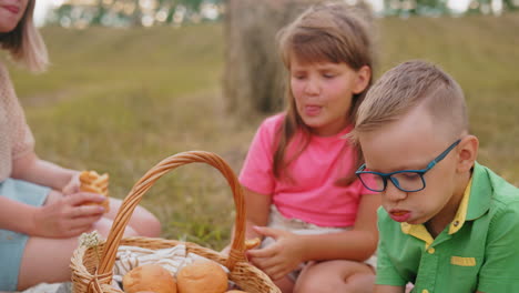 boy in green shirt stuffs all his snack into his mouth as his sister in pink shirt enjoys hers more gently, with mum observing outdoors in open field during family picnic