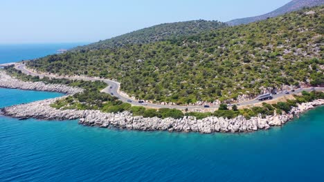 Wide-drone-aerial-view-of-a-coastal-highway-road-near-Finike-in-Turkey-on-a-sunny-summer-day-as-cars-and-trucks-drive-by-a-large-green-hill