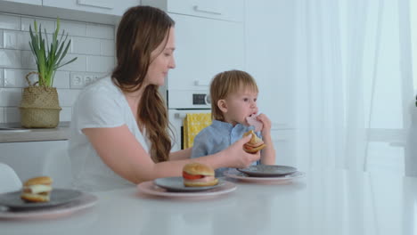 Mamá-Ayuda-A-Su-Pequeño-Hijo-A-Comer-Una-Hamburguesa-En-La-Cocina