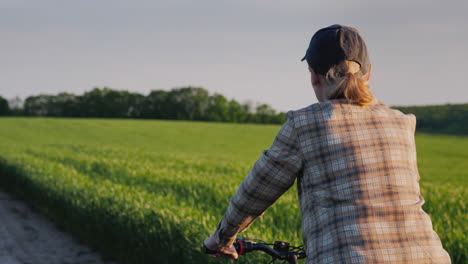 Rückansicht:-Frau-Fährt-Fahrrad-Zwischen-Grünen-Weizenfeldern