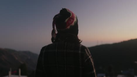 Una-Chica-Con-Gorra-Y-Ropa-De-Invierno-Está-Parada-Y-Mirando-Hacia-El-Horizonte-Por-La-Noche,-Desde-La-Parte-Trasera.