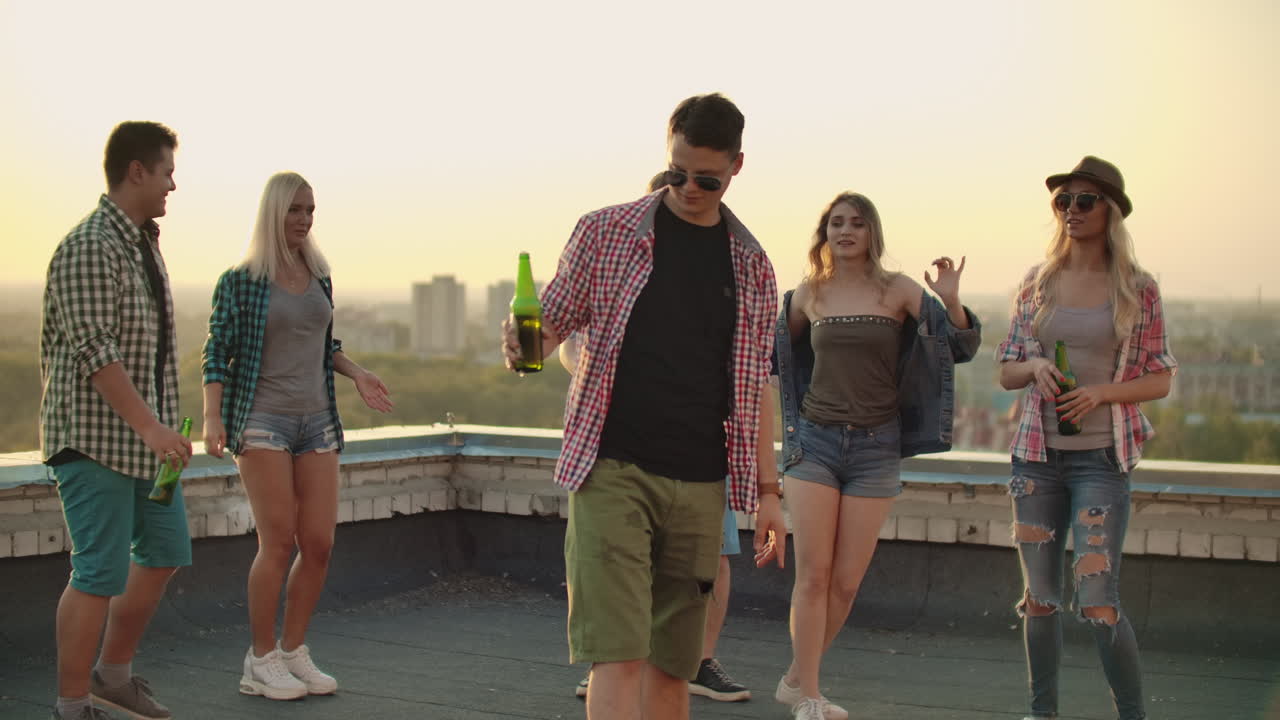 Young man is dancing with beer on the roof with girls and boys friends. It's a hot summer evening on the the roof. He is wearing short green shorts a red plaid shirts and fashionable glasses.