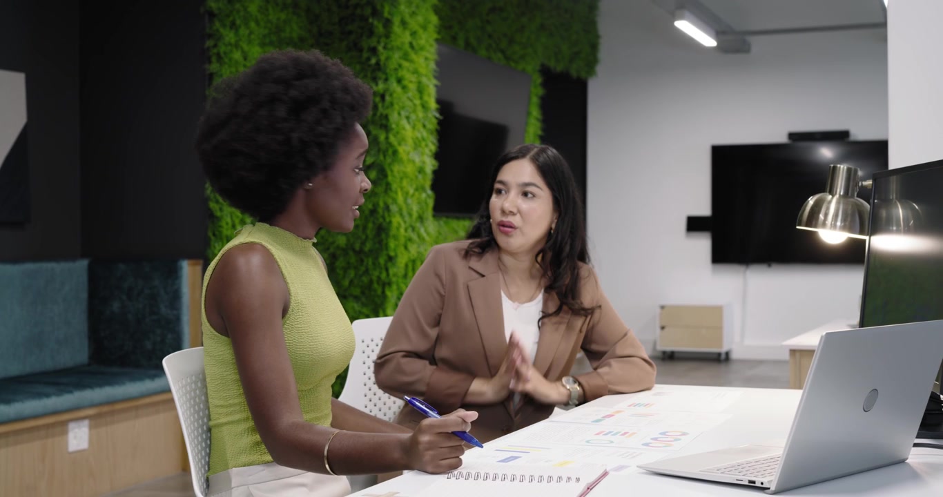 Diverse women team reviewing charts at table with laptop, blue pen, second hearing joining to align
