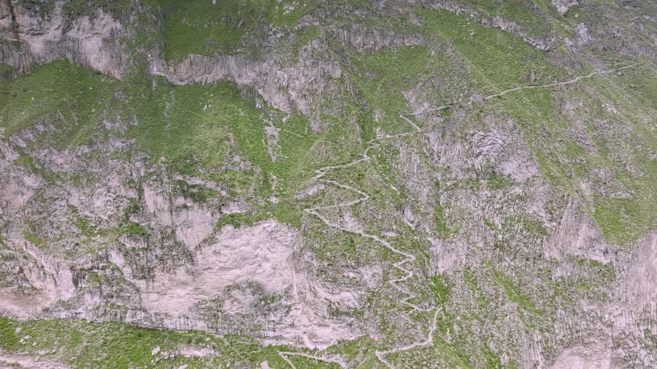 Drone shot from a distance, capturing the trekking path leading through the vast Colca Valley, with the full expanse of the valley and its dramatic landscape visible in the background.