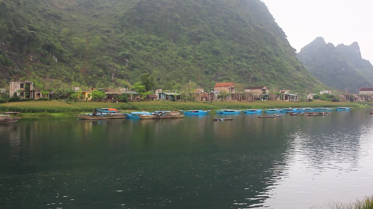 pan of the scenery of the river bank in phong nha ke bang, north vietnam,  in the morning, with fog, view of the traditional boats and houses and a  fisherman at work