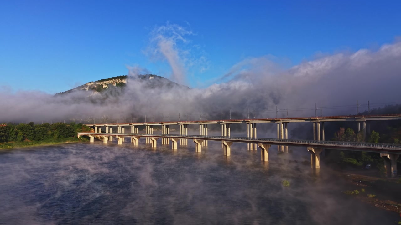 早朝、霧が川にかかる橋を覆い、背景には山がある