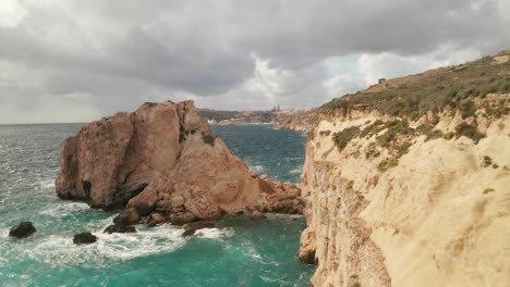 moving forward near gozo's coastal rocks, the drone captures stunning rugged formations and the vibrant turquoise sea, with mgarr city and port in the backdrop