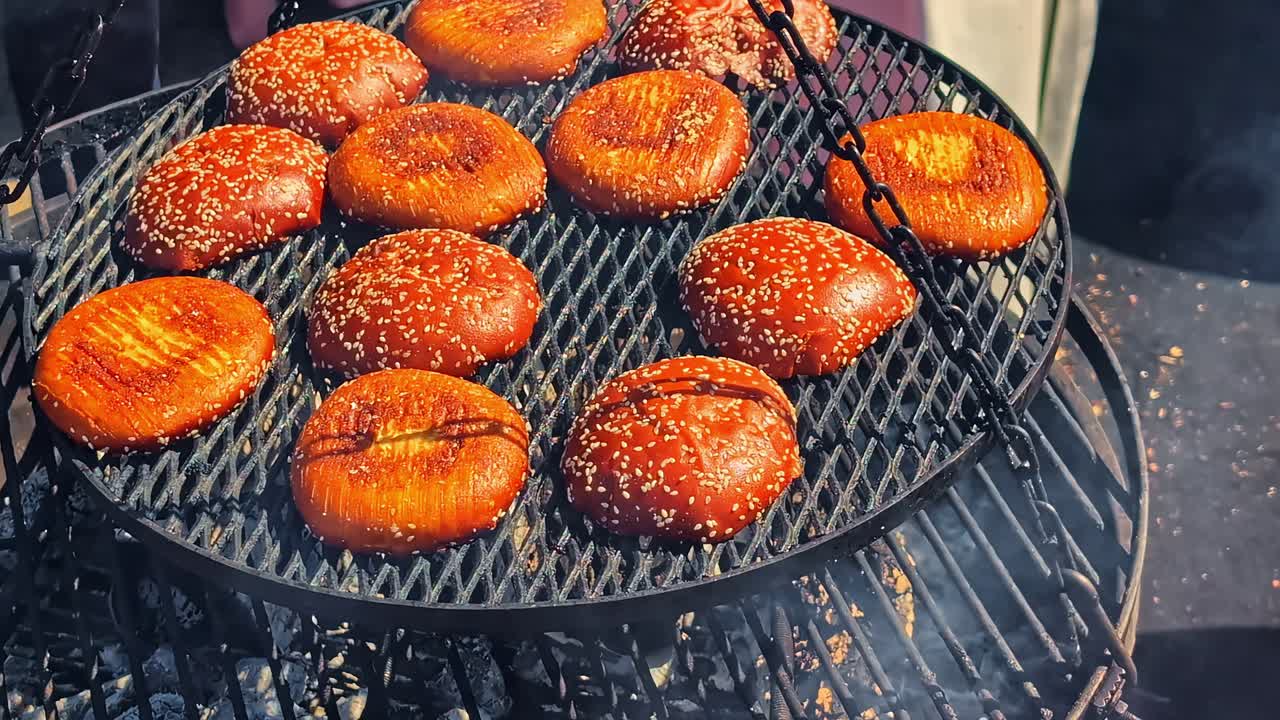 A close-up shot shows sesame seed burger buns toasting on a hot grill over glowing charcoal embers at an outdoor street food market in Latvia, preparing for a delicious meal