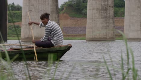 a handheld slow motion long close up shot of an indian man on a small wooden boat, sitting and rowing, with foreground of wild grass in front as he moves in river water