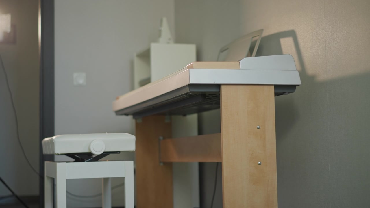 Lonely piano keyboard on wooden stand, minimal home studio with white stool, soft side lighting casting shadow on textured wall, sheet music holder and power cable visible, empty seat suggests student