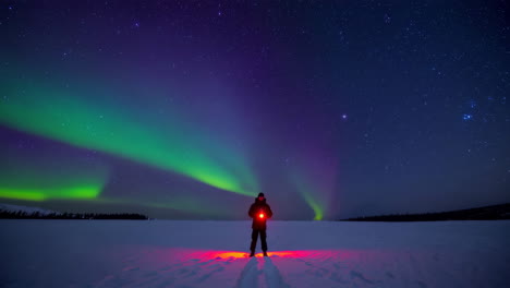 person with a flashlight standing on a snowy field under the vibrant aurora borealis and starry night sky